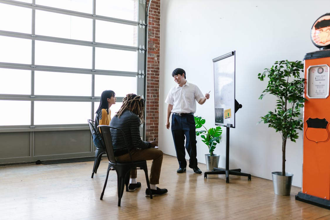 A man in a white shirt standing next to a flip chart, presenting to two seated individuals in a room with large windows and potted plants.