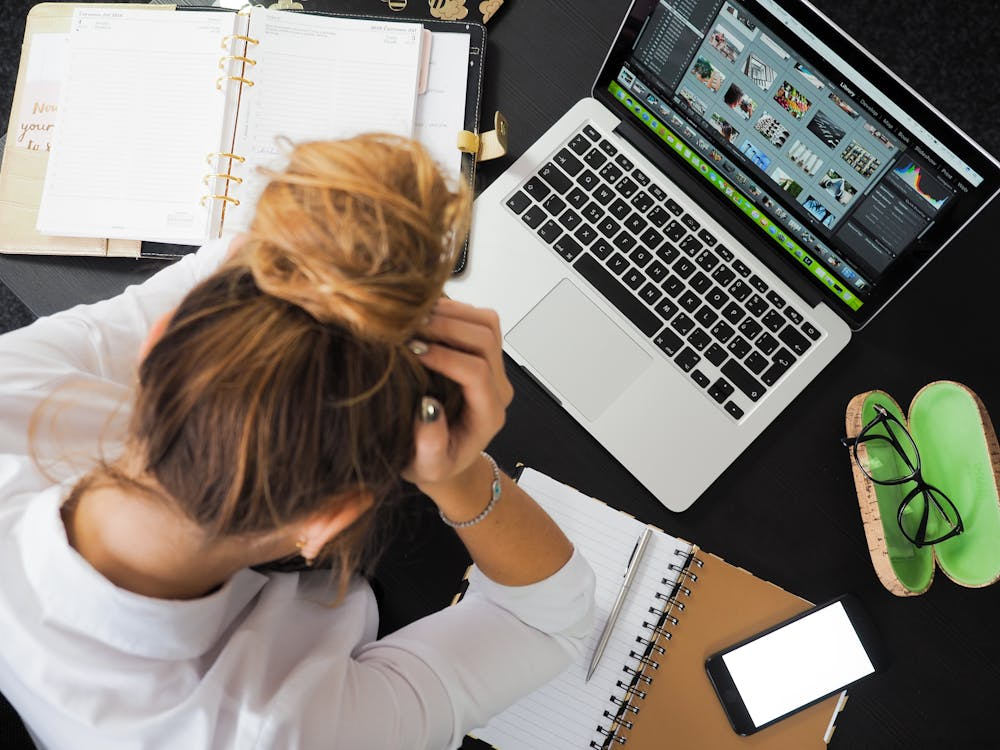 A woman with her hair in a bun sitting at a desk with her hands on her head, surrounded by a laptop, notebooks, smartphone, and eyeglasses.