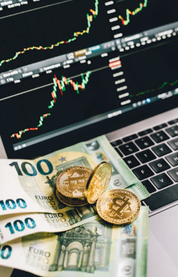 Gold-colored Bitcoin tokens and several 100 Euro banknotes placed on a laptop keyboard with trading charts displayed on the screen.