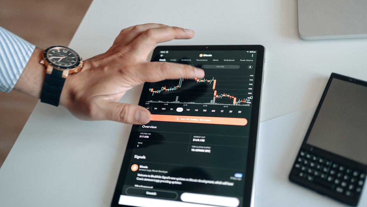 A person using a tablet displaying a candlestick chart of a cryptocurrency trading app, with one hand touching the screen.
