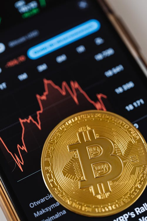 A large physical Bitcoin token placed on a smartphone screen displaying a red downward-trending financial chart.