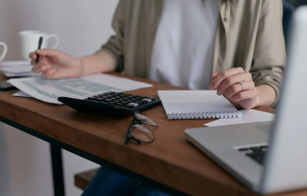 A person sitting at a wooden desk with a notebook, calculator, eyeglasses, documents, and a laptop, holding a pen in one hand.