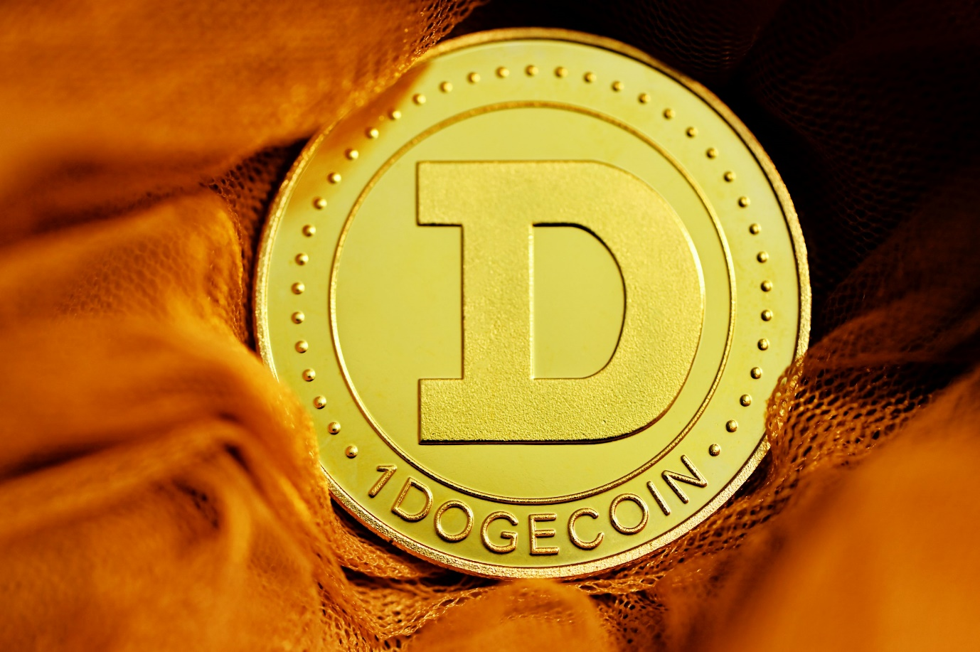 A gold-colored physical Dogecoin token marked “1 DOGECOIN” resting on an orange fabric background.