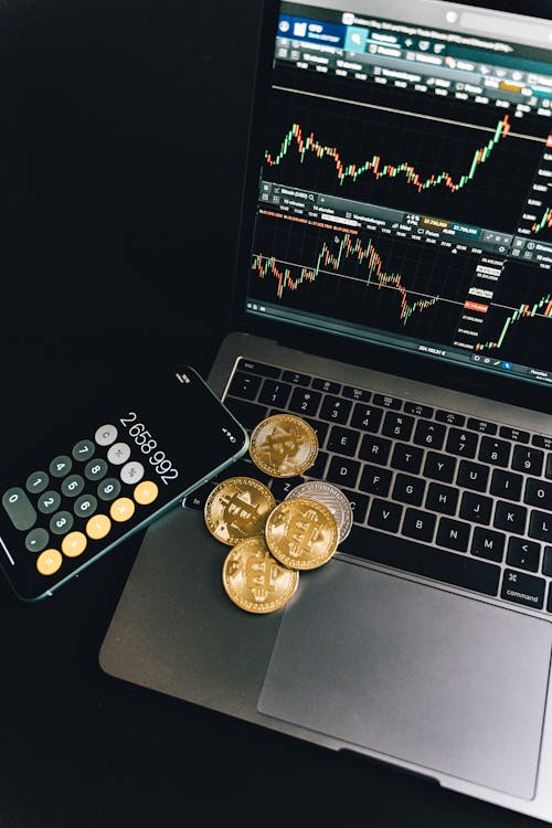 Gold and silver-colored Bitcoin tokens placed on a laptop keyboard next to a smartphone displaying a calculator app and a trading chart on the screen.