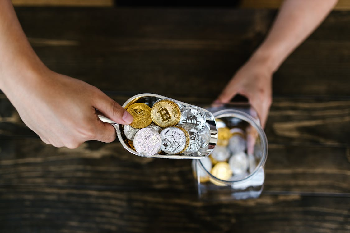 A person using a metal scoop to transfer silver and gold-colored physical cryptocurrency tokens into a glass jar on a wooden surface.