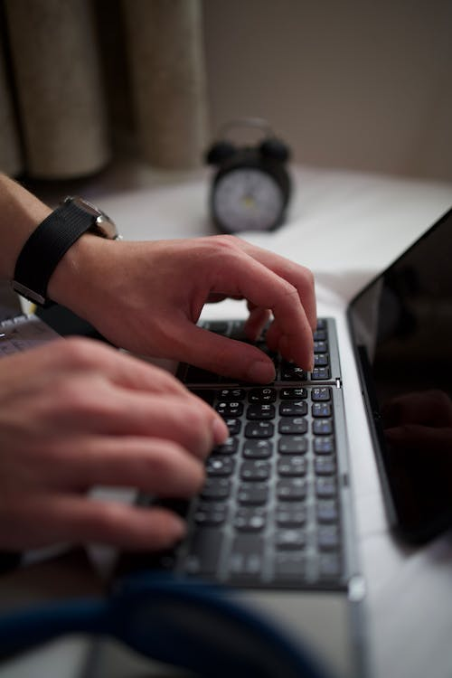 A person typing on a laptop keyboard with an analog alarm clock blurred in the background.