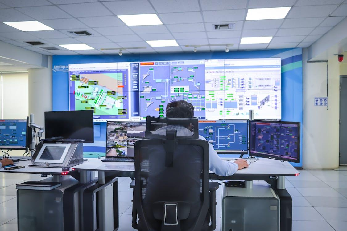 A person sitting in a control room in front of multiple computer monitors and large wall screens displaying system diagrams and data dashboards.