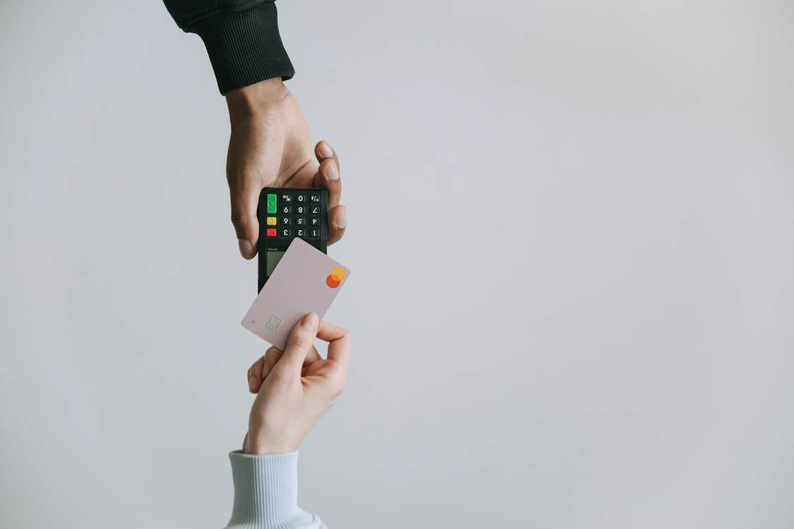 A person holding a pink credit card near a wireless card reader being held by another person, against a plain gray background.
