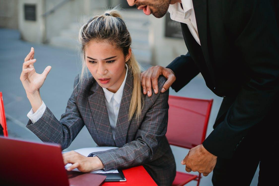 A woman in a plaid blazer sitting at a red table looking frustrated at her laptop, while a man stands beside her with his hand on her shoulder.