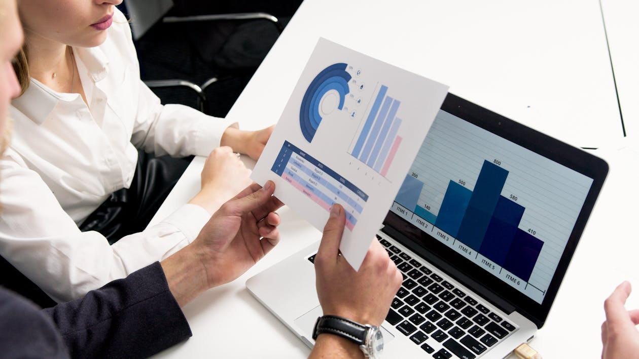 Two people reviewing a printed chart with bar graphs and circular data visuals, while a laptop on the table displays a blue bar graph.