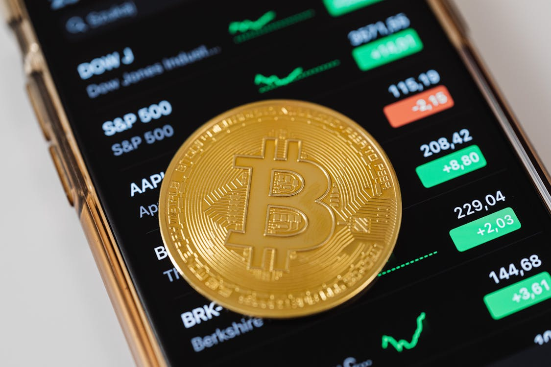 A physical gold-colored Bitcoin placed on top of a smartphone screen displaying a stock market app with various indices and price movements.