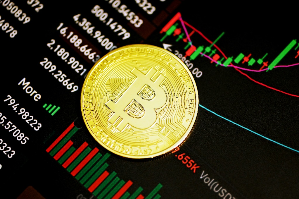 A golden Bitcoin coin resting on a dark screen displaying real-time cryptocurrency trading charts, data, and price fluctuations.